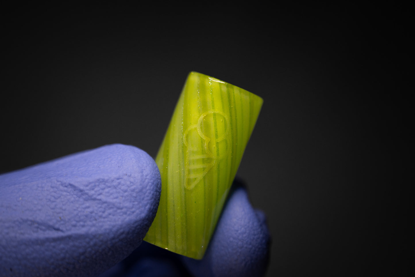 A gloved hand holds a small, cylindrical GREEN 12MM STRAIGHT by CONEZ against a dark background, featuring an engraved design of a bee with wings and antennae.