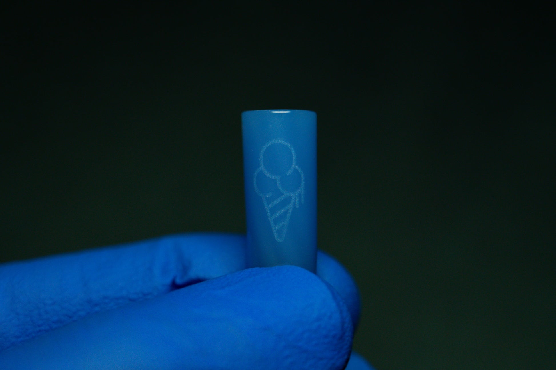 A gloved hand grips a BLUE CONEZ by CONEZ, a small cylindrical item laser-engraved with a two-scoop ice cream cone. Both the object and glove are blue, set against a dark green backdrop.