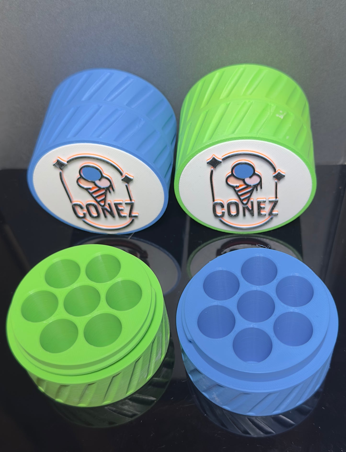 Two colorful cylindrical TRAVEL CASE gadgets from CONEZ are displayed—one blue, one green—each with compartments on top. Both have a white lid with an ice cream cone logo and the word CONEZ, placed on a reflective surface.