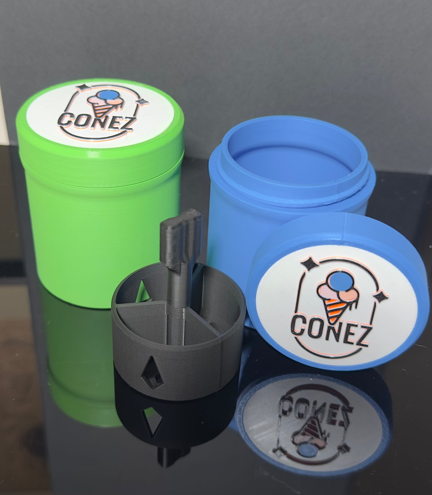 On a reflective surface sit two plastic containers by CONEZ. The green ISO DUNK TANK is closed, while the blue one is open, revealing a black cylindrical insert with side openings. These containers simplify cleaning with isopropyl alcohol tips and are ideal for use as dunk tanks.